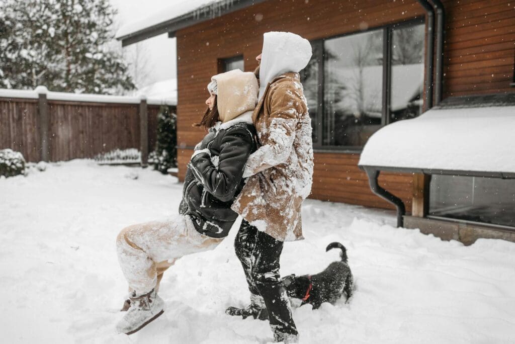 A couple dressed in winter clothes playing in the snow with their pet dog outside a house.