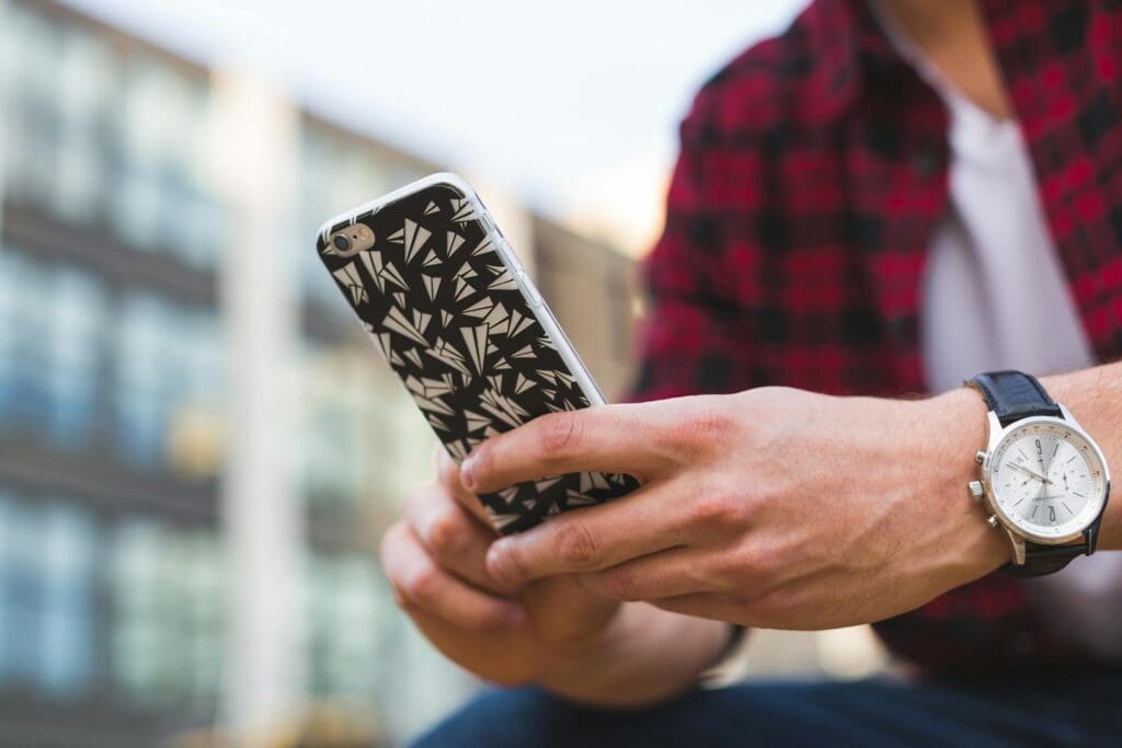 Close-up of a man holding a smartphone with a patterned case, wearing a wristwatch outdoors.