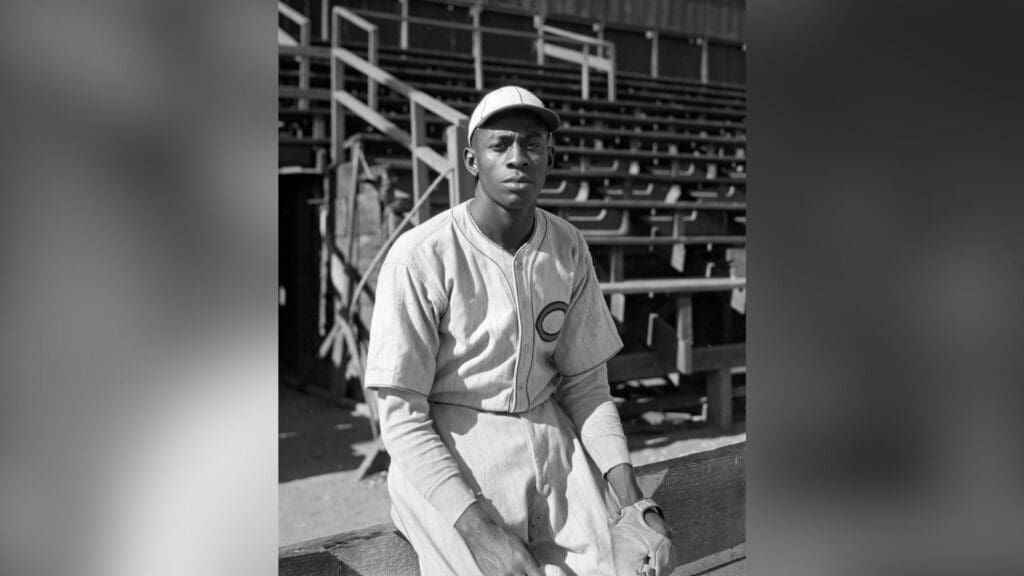 Baseball pitcher Satchel Paige seated next to bleachers. The location may be Thomas Jefferson High School at Hopner and 41st St., Los Angeles.
